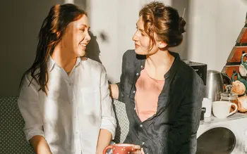 Moderne Wohnküche in sanften Grüntönen mit rundem Esstisch und großen Fenstern, daneben zwei Frauen, die auf einer Küchenbank zusammensitzen und bei Kaffee lächeln. Kombination aus Designküche und warmem Miteinander.